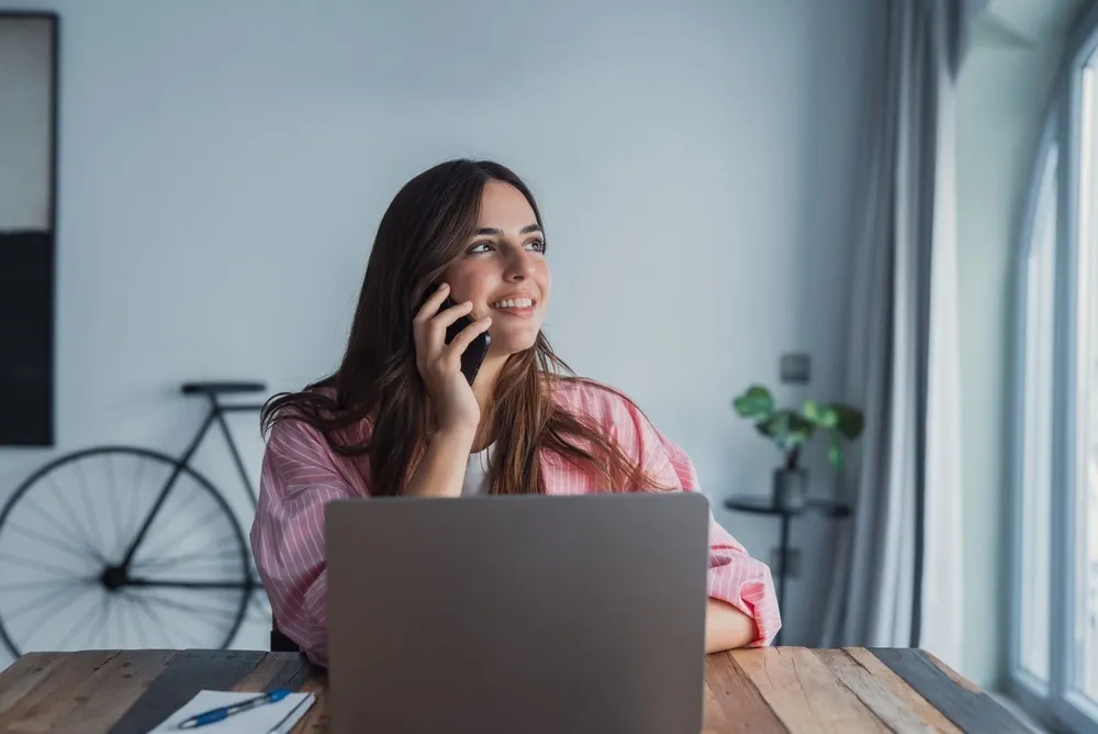 Smiling woman sits at a wooden table with a laptop, talking on her phone
